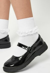 Svart lackläder Mary Jane-skor med rund tå och rem. Bärs med vita volangiga ankelsockor. Strukturerad svart gummisula.