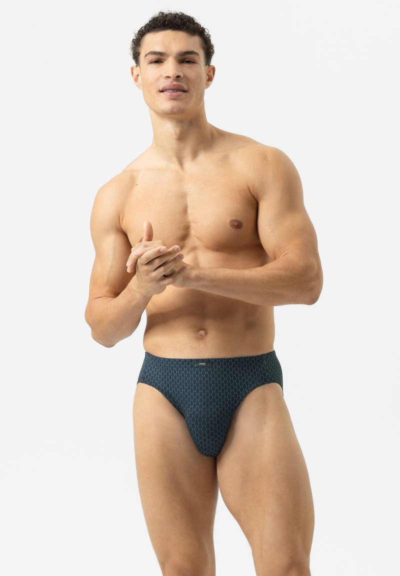 Spierachtige man die staat en zijn handen vouwt, gekleed in donkere patroon ondergoed tegen een eenvoudige witte achtergrond.