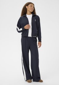 Femme portant un survêtement bleu marine avec des rayures blanches, un t-shirt blanc et des chaussures marron, se tenant devant un fond blanc uni.