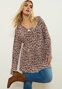 Långärmad blommig tunika med V-ringning i krämfärg, med ett mönster av maroon- och rostfärgade blommor, samt vågig nederkant. Stylist ihop med blå jeans och bruna stövlar.