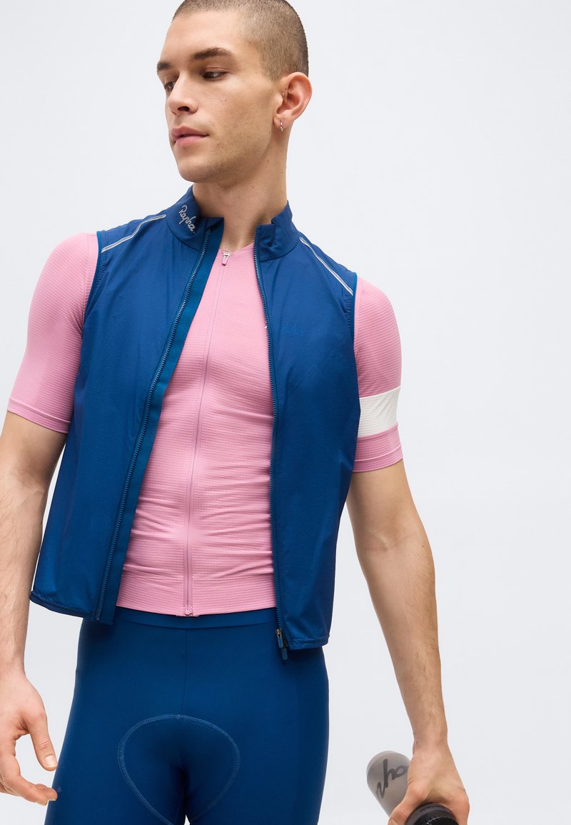 Male cyclist wearing a pink cycling jersey and blue vest, holding a bicycle handlebar, looking to the side against a white background.