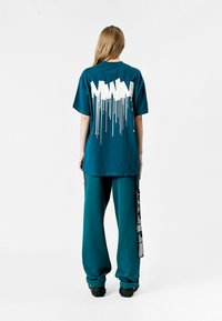 Camiseta de algodón de gran tamaño en color verde azulado con un diseño gráfico blanco y efecto de goteo en la parte posterior. Combinada con pantalones sueltos a juego y calzado negro.