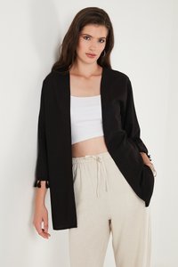 Cardigan negro abierto con mangas de tres cuartos, que presenta acentos de borlas en los puños; combinado con una blusa blanca corta y pantalones beige claros.