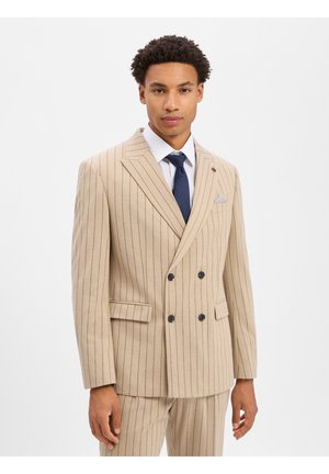 Giovane uomo che indossa un abito beige a righe sottili doppiopetto con camicia bianca e cravatta blu navy, posando su uno sfondo bianco semplice.