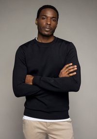 Sudadera negra con cuello redondo y mangas largas. Textura suave y diseño ajustado. Camiseta blanca visible en el dobladillo.
