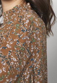 Brown floral blouse featuring a delicate pattern of white and blue flowers, green leaves, and a high ruffled neckline. Soft fabric texture.