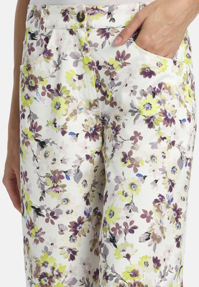 Pantalon blanc avec un motif floral jaune, violet et vert, fermeture à boutons, poches avant, une main partiellement dans une poche.