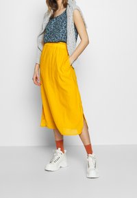 Cardigan gris clair en maille, débardeur bleu à fleurs, jupe midi jaune avec fentes latérales, et baskets blanches épaisses avec des chaussettes orange.