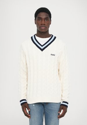 Cremefarbener Kabelstrickpullover mit navyblauen und hellblauen Streifen im V-Ausschnitt. Mit gerippten Bündchen, die mit Streifen verziert sind, und einem gestickten Logodetail.