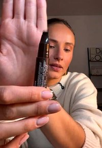 Mujer sosteniendo un lápiz de cejas negro con punta multipunta cerca de la cámara, vistiendo un suéter claro en el interior.