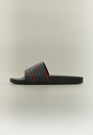 Schwarze Slidesandale mit strukturierter Sohle und Riemen, der das wiederholte "HUGO"-Logo in schwarzer und roter Schrift zeigt.