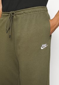 Close-up van een persoon die olijfgroene Nike-sweatpants draagt met een trekkoord in de taille en een hand in de zak tegen een witte achtergrond.