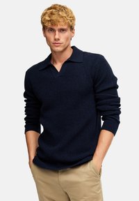 Maglione lavorato a maglia blu navy con scollo a V e design con colletto. Presenta maniche lunghe e un tessuto texturizzato. Abbinato a pantaloni beige chiaro.