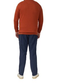 Man in een roestrood gebreid vest, marineblauwe broek en witte sneakers, staand met zijn rug naar de camera tegen een witte achtergrond.
