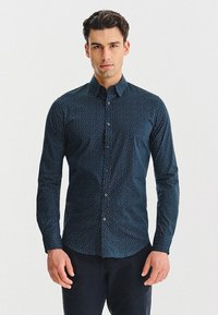 Mężczyzna w ciemnoniebieskiej koszuli z długim rękawem zapinanej na guziki, w mały biały wzór kropek, stojący na tle białego tła.