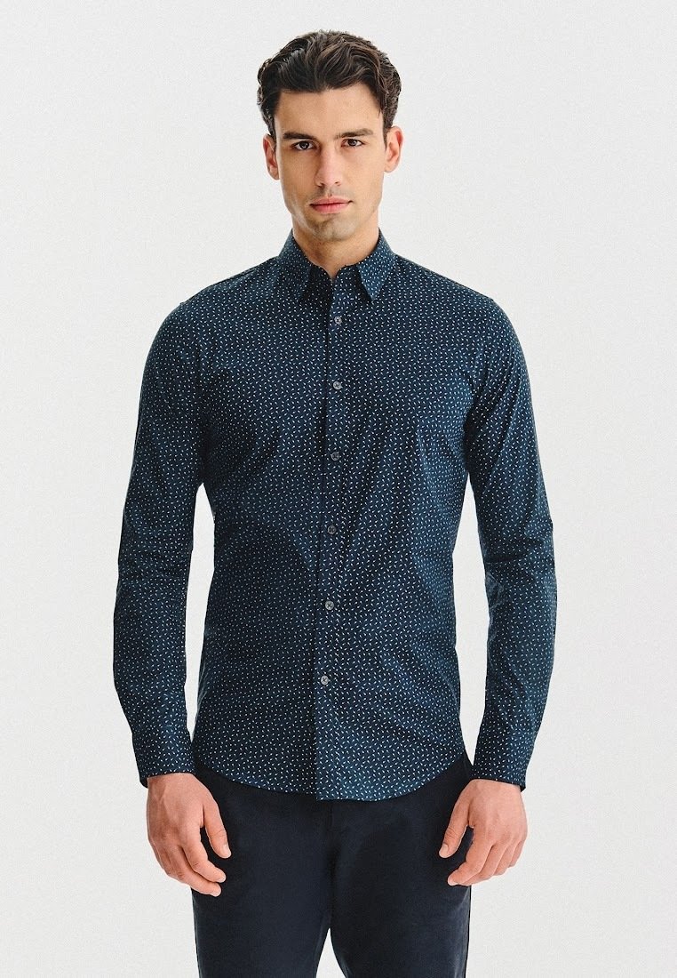 Mężczyzna w ciemnoniebieskiej koszuli z długim rękawem zapinanej na guziki, w mały biały wzór kropek, stojący na tle białego tła.