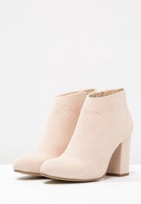 Botines beige de ante suave con punta redondeada y tacón alto de bloque. Presentan una cremallera lateral para facilitar su uso y un diseño curvado y suave.