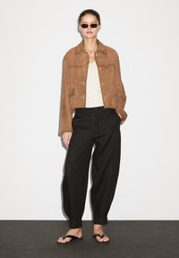 Brun ruskind cropped jakke, hvid top, sorte vidde bukser og sorte flip-flops. Modellen bærer runde solbriller. Simpelt baggrund.