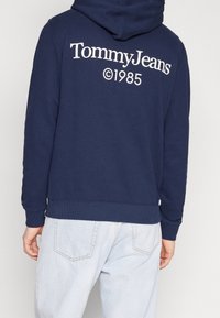 Námořnická modrá mikina s kapucí se velkým bílým logem "Tommy Jeans ©1985" na zadní straně. Obsahuje šňůrku na stažení kapuce a žebrované manžety.