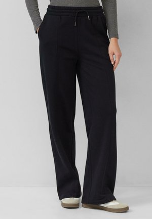 Frau trägt schwarze weite Hosen mit vorderen Nähten, ein graues Langarmoberteil und beige Sneakers vor einem schlichten Hintergrund.