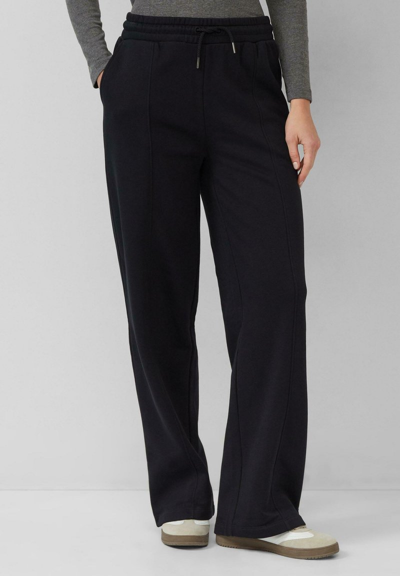 Kobieta w czarnych szerokich spodniach z przednimi szwami, szarej bluzce z długim rękawem i beżowych trampkach na jednolitym tle.