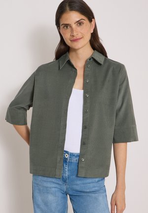 Jeune femme aux cheveux bruns et aux taches de rousseur portant une chemise vert foncé déboutonnée sur un haut blanc et un jean bleu, debout devant un fond uni.