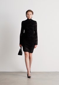 Μαύρο φόρεμα από δαντέλα με μακριά μανίκια, ψηλό λαιμό και εφαρμοστή σιλουέτα. Το μοντέλο κρατά μια μικρή μαύρη τσάντα και φορά μαύρες γόβες. Λείος υφασμάτινος λαμπερός.