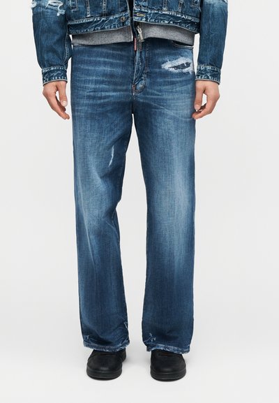 Άτομο που φορά μπλε τζιν με φθαρμένο στυλ και τζιν μπουφάν, στέκεται με μαύρα παπούτσια σε απλό φόντο.