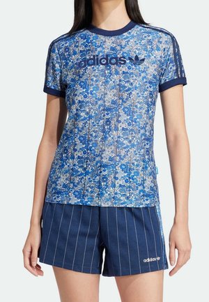 Kobieta ubrana w niebieską kwiecistą koszulkę Adidas z granatowym wykończeniem oraz granatowe krótkie spodenki Adidas w cienkie paski z logo na lewej nodze.