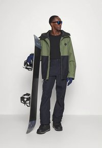 En snowboarder har på seg en grønn og marineblå vanntett jakke, svart termisk skjorte, marineblå bukser, blå hansker og svarte støvler, og holder et svart snowboard.