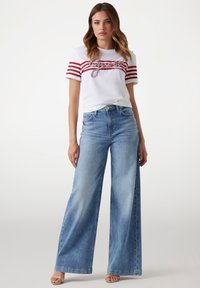 Witte t-shirt met rode strepen en grafische tekst, gecombineerd met lichte spijkerbroek met wijde pijpen. De outfit heeft casual schoeisel.