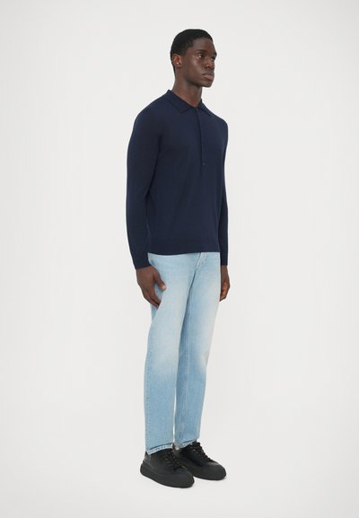 Uomo in camicia polo a maniche lunghe blu navy, jeans azzurri e sneakers nere su uno sfondo bianco neutro.