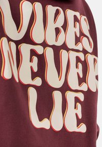 Bordeaux sweatshirt met gedurfde, golvende witte tekst met geel-oranje accenten, met de tekst "VIBES NEVER LIE". Zachte textuur, stof van gemiddelde gewicht.