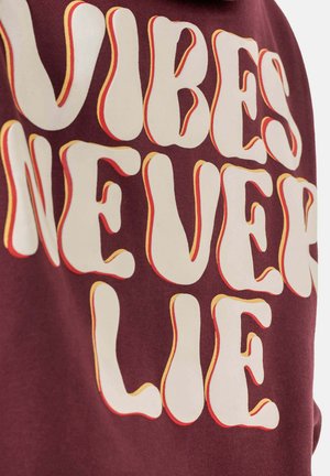 Bordeauxfarbenes Sweatshirt mit fettgedrucktem, gewundenem weißen Text und gelb-orangefarbenen Akzenten, der "VIBES NEVER LIE" sagt. Weiche Textur, mittelschwerer Stoff.