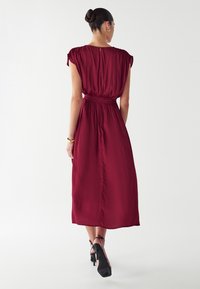 Vestido midi color burdeos con mangas cortas, cinturilla elástica y textura suave. Combinado con tacones negros de tiras y accesorios dorados sencillos.