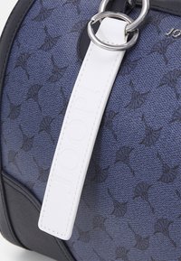 Sac à main bleu à motifs avec un corps texturé, des accents noirs et une étiquette logo blanche attachée par un anneau en argent.