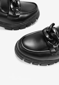 Zapatos de cuero negro con un acabado brillante, que presentan una suela gruesa y un gran detalle de cadena en la parte superior. Textura suave con costuras en contraste.