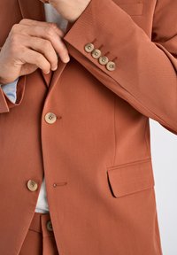 Blazer de color óxido con una textura suave, que cuenta con cuatro botones, un único bolsillo en el pecho y costuras de contraste en el puño.