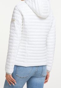 Kobieta ubrana w dopasowaną białą pikowaną kurtkę z kapturem oraz jasnoniebieskie dżinsy, pokazana od tyłu i z boku.