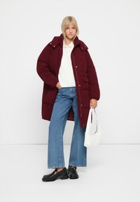 Bordeaux pufferjakke med høj krave, knaplukning foran og påsætningslommer, parret med en hvid sweater, blå vide jeans og sorte sko.