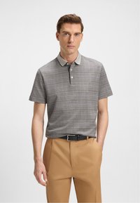Hombre con una camiseta polo de color gris con patrón, cuello gris claro, pantalones color camel y cinturón negro, de pie con una mano en el bolsillo.