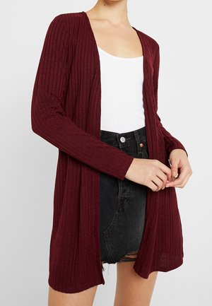 Femme portant un cardigan bordeaux côtelé à manches longues et ouvert sur le devant, par-dessus un débardeur blanc et un short en jean noir taille haute.