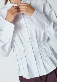 Chemise blanche à boutons, à manches longues, avec un design froncé à la taille et une texture lisse. Dotée d'un col classique et de boutons en nacre.