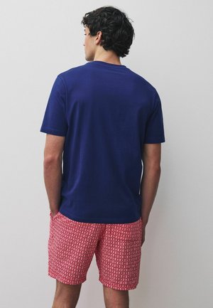 Mann mit dem Rücken zur Kamera, trägt ein schlichtes dunkelblaues kurzärmeliges Shirt und rote gemusterte Shorts, die Hände in den Taschen, vor einem einfarbigen Hintergrund.