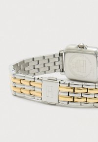 Kovový náramok na hodinky so striebornými a zlatými článkami a štvorcovou sponou s vygravírovaným logom, pripevnený k štvorcovému puzdru hodiniek.
