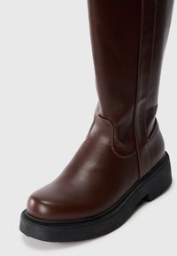 Bota de cuero marrón hasta la rodilla con punta redonda, costuras en contraste y una suela de goma negra gruesa. Textura suave y detalles de diseño mínimos.