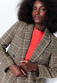 Blazer à carreaux brun avec des épaules structurées et deux boutons à l'avant, superposé sur un haut orange vif. Texture douce, lignes définies.