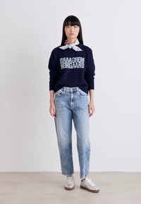 Felpa navy con logo "Nørgaard", jeans a gamba larga di colore azzurro chiaro e sneakers grigie. Modello che indossa una sciarpa bianca e rossa a fantasia.