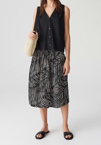 Musta ilmanhihaisella paidalla, jossa on napit, yhdistetty musta ja kerma valkoinen kuvioitu hame. Mallilla on kädessään kudottu beige laukku ja jalassa mustat liukulapasandalit.