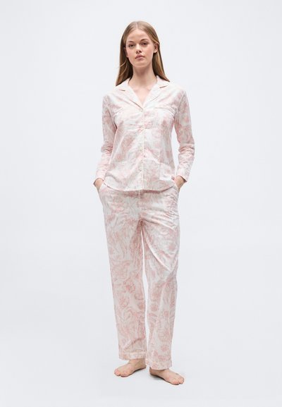 Νεαρή γυναίκα στέκεται ξυπόλυτη φορώντας ανοιχτό ροζ πουκάμισο πιτζάμας με λουλουδάτο μοτίβο και αντίστοιχο φαρδύ παντελόνι.